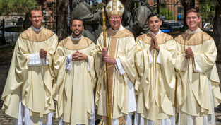 Five Catholic clergy in cream vestments pose outdoors, smiling, with a bishop holding a staff before bronze statues.