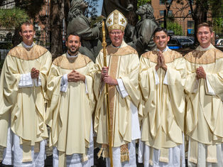 Five Catholic clergy in cream vestments pose outdoors, smiling, with a bishop holding a staff before bronze statues.