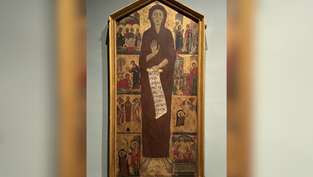 Tall framed medieval icon of a haloed saint holding a scroll with Latin text, surrounded by small biblical scenes in a museum gallery