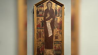 Tall framed medieval icon of a haloed saint holding a scroll with Latin text, surrounded by small biblical scenes in a museum gallery
