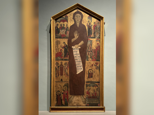 Tall framed medieval icon of a haloed saint holding a scroll with Latin text, surrounded by small biblical scenes in a museum gallery