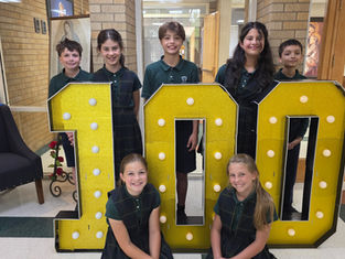 Six smiling students in green uniforms pose behind a glittery gold 100 sign in a school hallway; Saint Joseph Middle School poster nearby.
