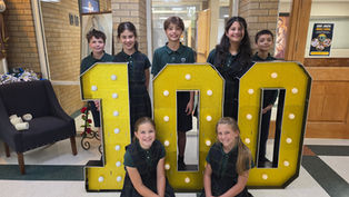 Six smiling students in green uniforms pose behind a glittery gold 100 sign in a school hallway; Saint Joseph Middle School poster nearby.