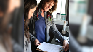 Two people in a clinic examine a clipboard with papers. One wears a stethoscope, smiling. Background shows medical supplies by a window.