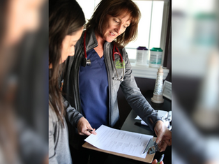 Two people in a clinic examine a clipboard with papers. One wears a stethoscope, smiling. Background shows medical supplies by a window.