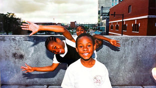 Three smiling children pose playfully on a concrete ledge with arms outstretched. Urban skyline and brick buildings in the background.