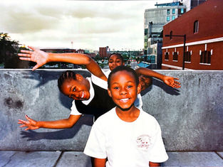 Three smiling children pose playfully on a concrete ledge with arms outstretched. Urban skyline and brick buildings in the background.