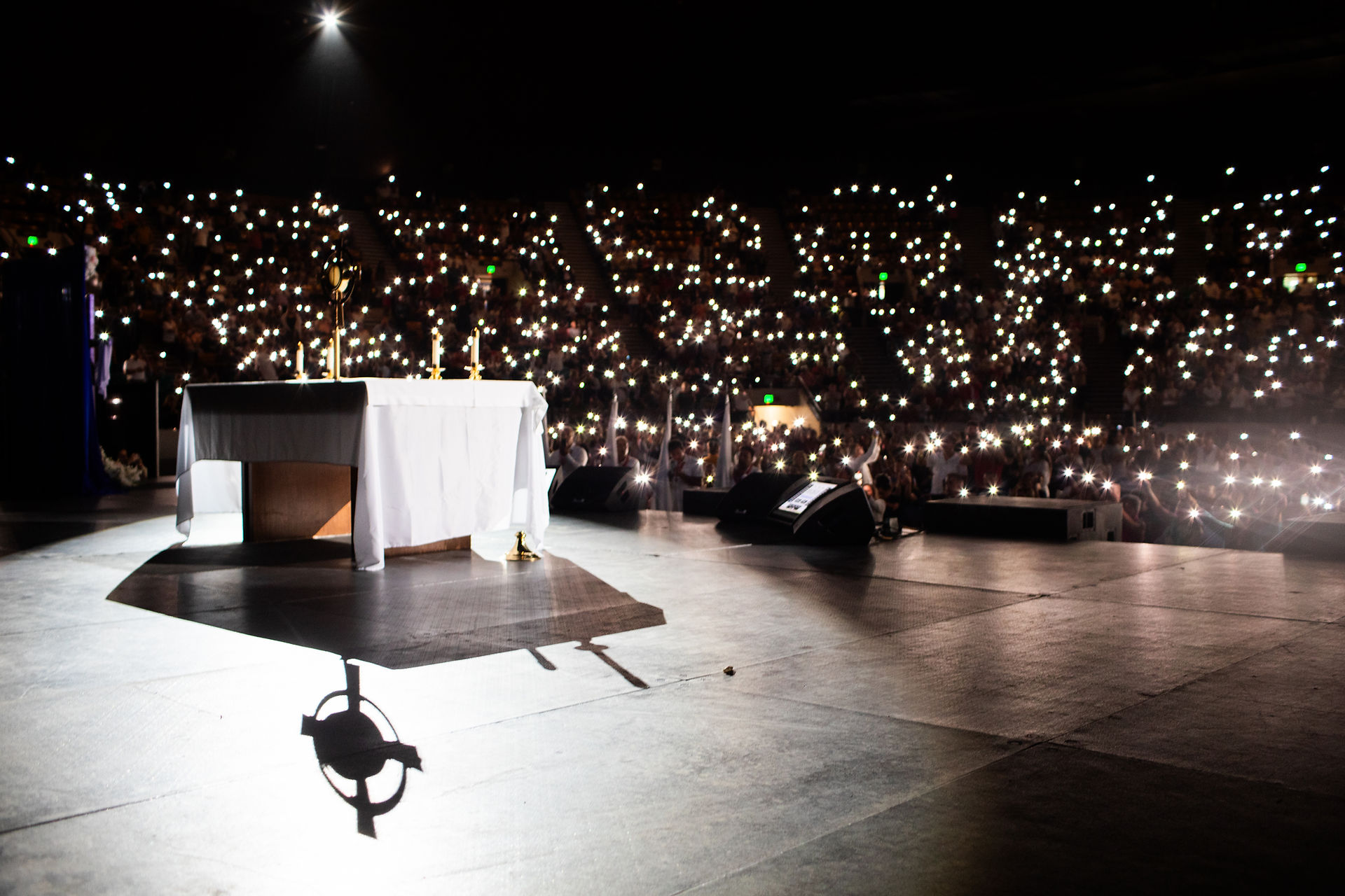 White-clothed altar with candles on stage. Audience in background holding lit phones, creating a sea of lights. Dark, atmospheric setting.