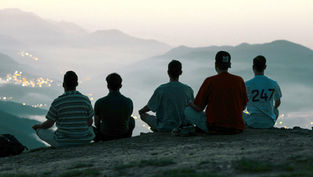 Five people sit on a hill overlooking a misty valley at dusk. One wears a "247" shirt. City lights twinkle below, creating a tranquil mood.