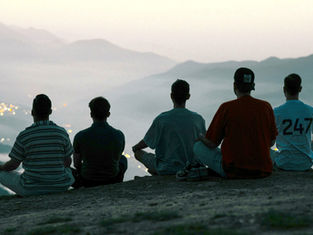Five people sit on a hill overlooking a misty valley at dusk. One wears a "247" shirt. City lights twinkle below, creating a tranquil mood.