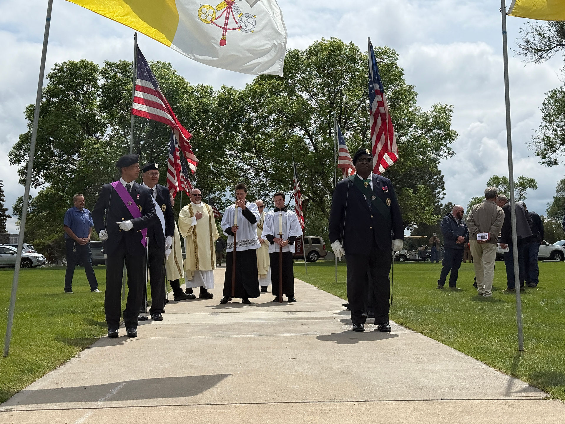 People in ceremonial attire walk on a path with flags in a grassy area. Some wear religious robes, others, dark suits. Trees and parked cars in the background.