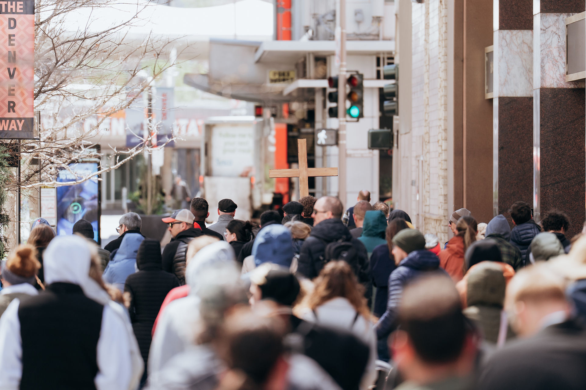 A crowd walks through a downtown street, following a wooden cross during a public religious procession.