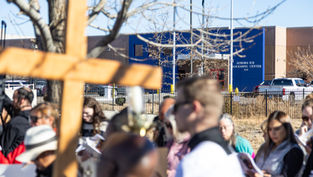 A group gathers near a tree holding a large wooden cross at Aurora ICE Processing Center. People read papers; mood is solemn. Blue sky above.