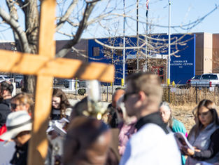 A group gathers near a tree holding a large wooden cross at Aurora ICE Processing Center. People read papers; mood is solemn. Blue sky above.