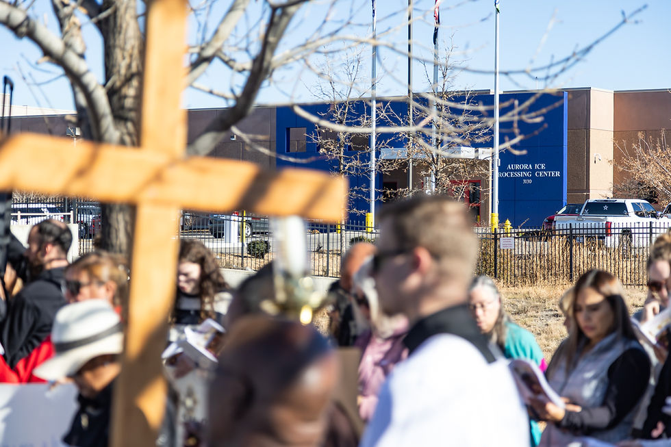 A group gathers near a tree holding a large wooden cross at Aurora ICE Processing Center. People read papers; mood is solemn. Blue sky above.