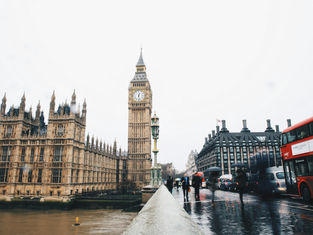 View of Big Ben and the Houses of Parliament in London. Red double-decker buses and people with umbrellas on wet street. Overcast sky.
