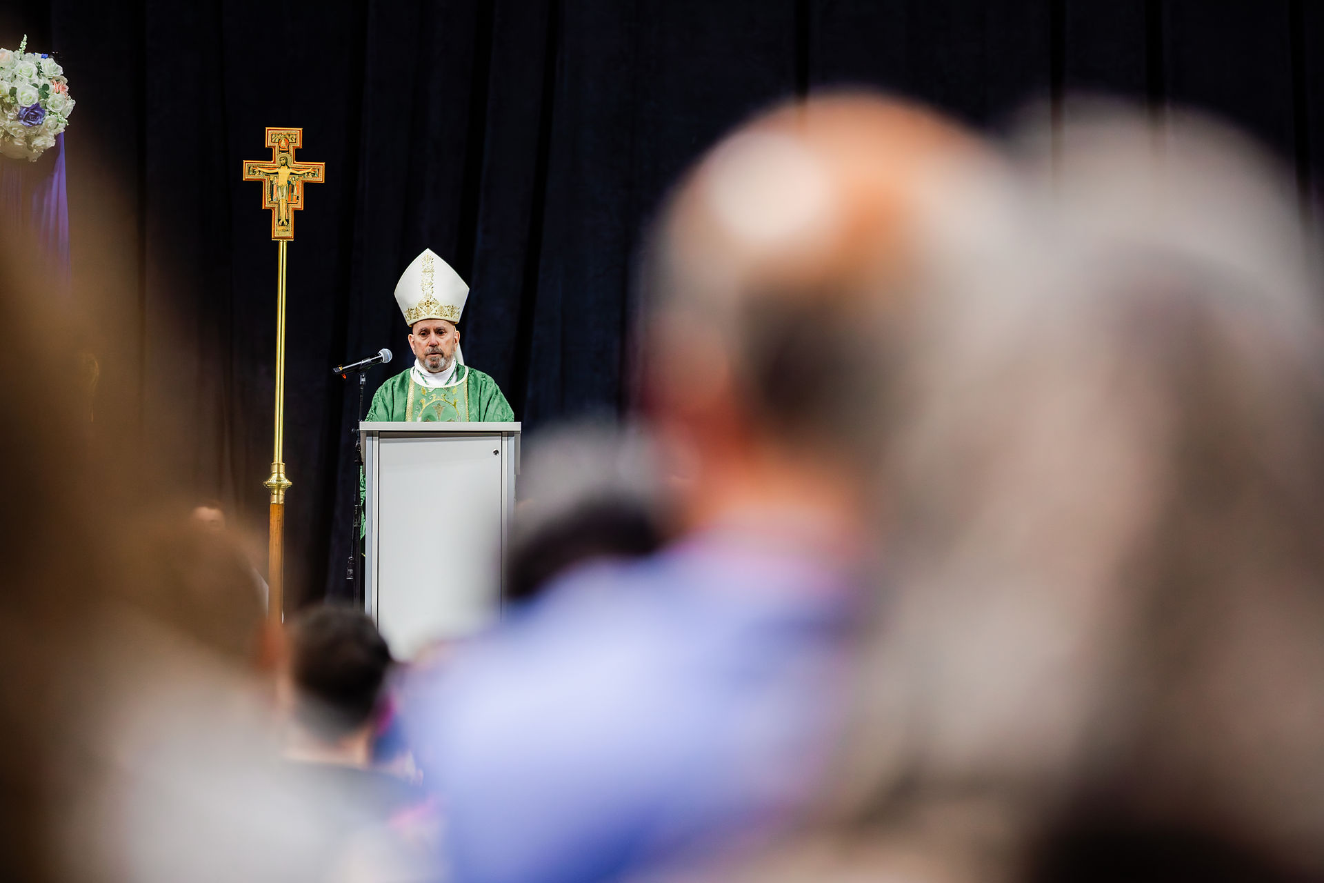 Bishop in green vestments speaks at a podium with a cross beside him. Dark background, blurred crowd in foreground. Solemn atmosphere.