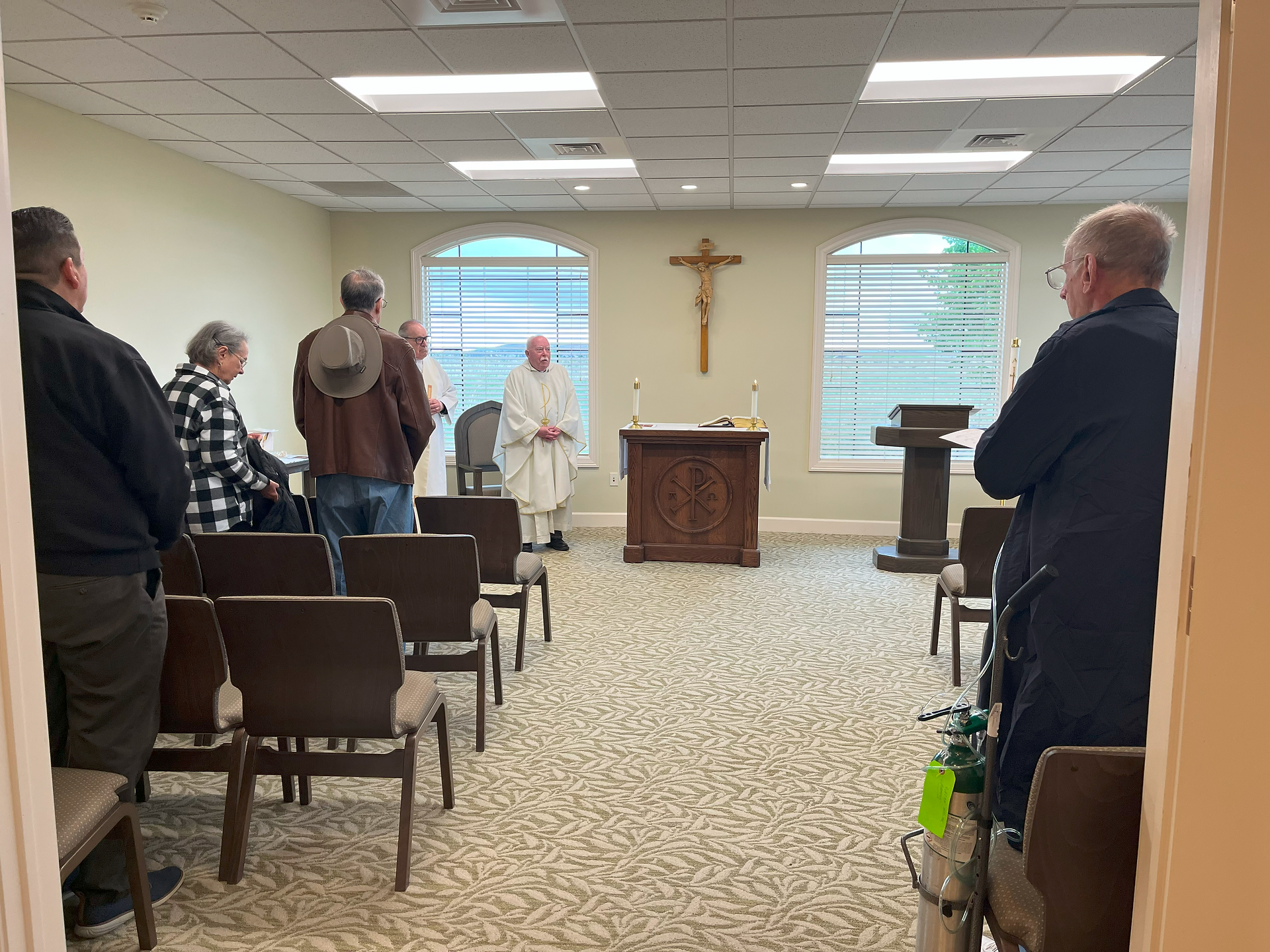 A small chapel with people standing, facing a priest dressed in white robes near an altar. Cross on wall, calm atmosphere, indoor setting.