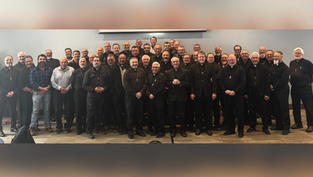 Group of men, mostly in black attire, smiling in a conference room with a projector screen behind. Warm, professional atmosphere.