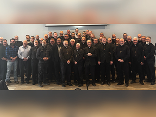 Group of men, mostly in black attire, smiling in a conference room with a projector screen behind. Warm, professional atmosphere.