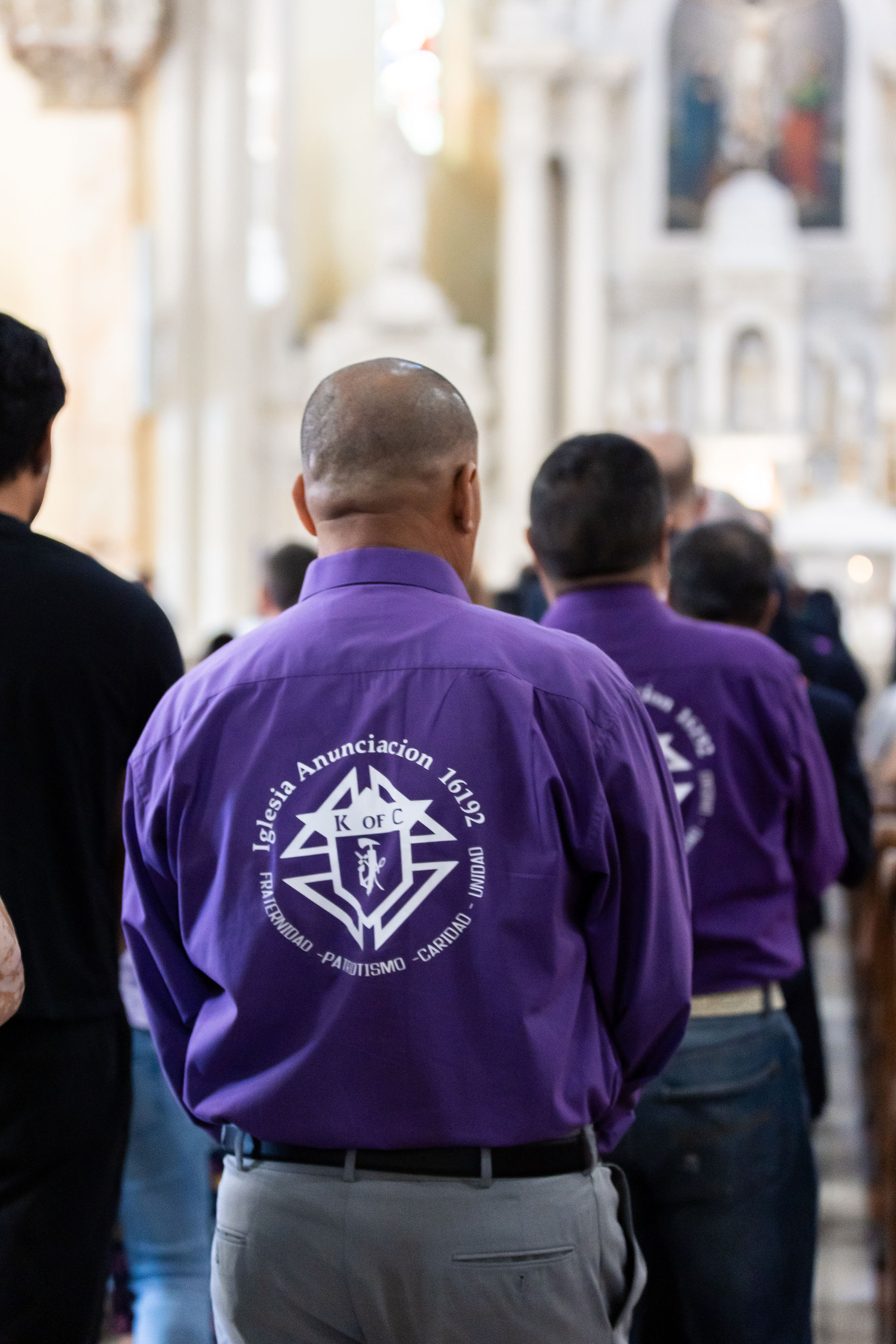 Men in purple shirts with church logos stand in a cathedral. The text reads "Iglesia Anunciacion." The atmosphere is solemn.