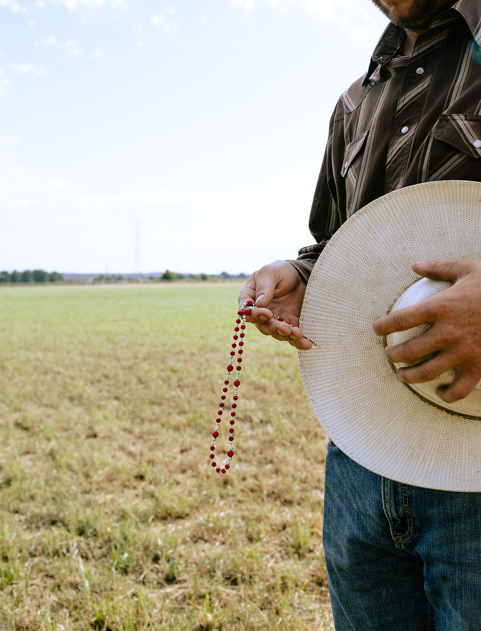 Man holding a straw hat and red rosary in a grassy field, wearing a striped brown shirt and jeans. Sky is partly cloudy. Calm atmosphere.