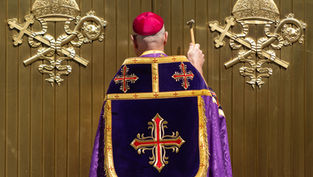 A person in religious attire, holding a hammer, faces a gold door with ornate emblems, wearing a purple and red embroidered robe.