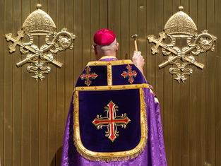 A person in religious attire, holding a hammer, faces a gold door with ornate emblems, wearing a purple and red embroidered robe.