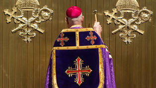 A person in religious attire, holding a hammer, faces a gold door with ornate emblems, wearing a purple and red embroidered robe.