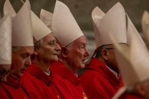 Clergy in white mitres and red robes sit solemnly in a dimly lit setting, focusing intently, with no visible text.