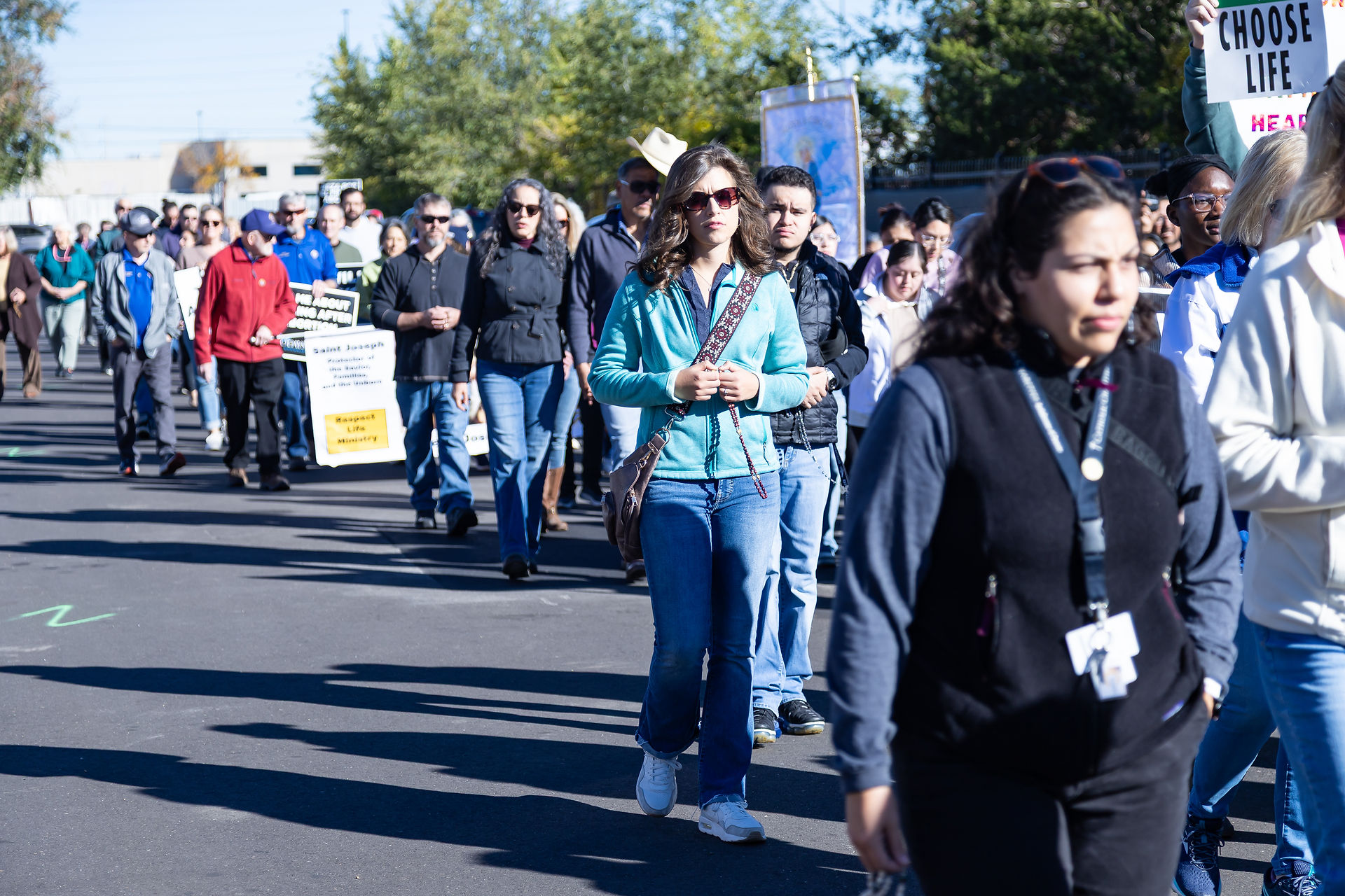 A crowd walks in a peaceful protest, holding signs that read "Choose Life." The setting is outdoors with trees and a clear sky.