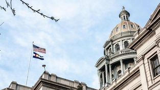 The U.S. and Colorado flags fly atop a historic domed building against a clear sky, framed by bare branches, conveying a serene mood.