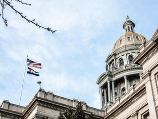 The U.S. and Colorado flags fly atop a historic domed building against a clear sky, framed by bare branches, conveying a serene mood.
