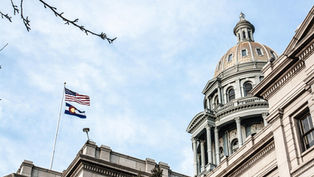 The U.S. and Colorado flags fly atop a historic domed building against a clear sky, framed by bare branches, conveying a serene mood.