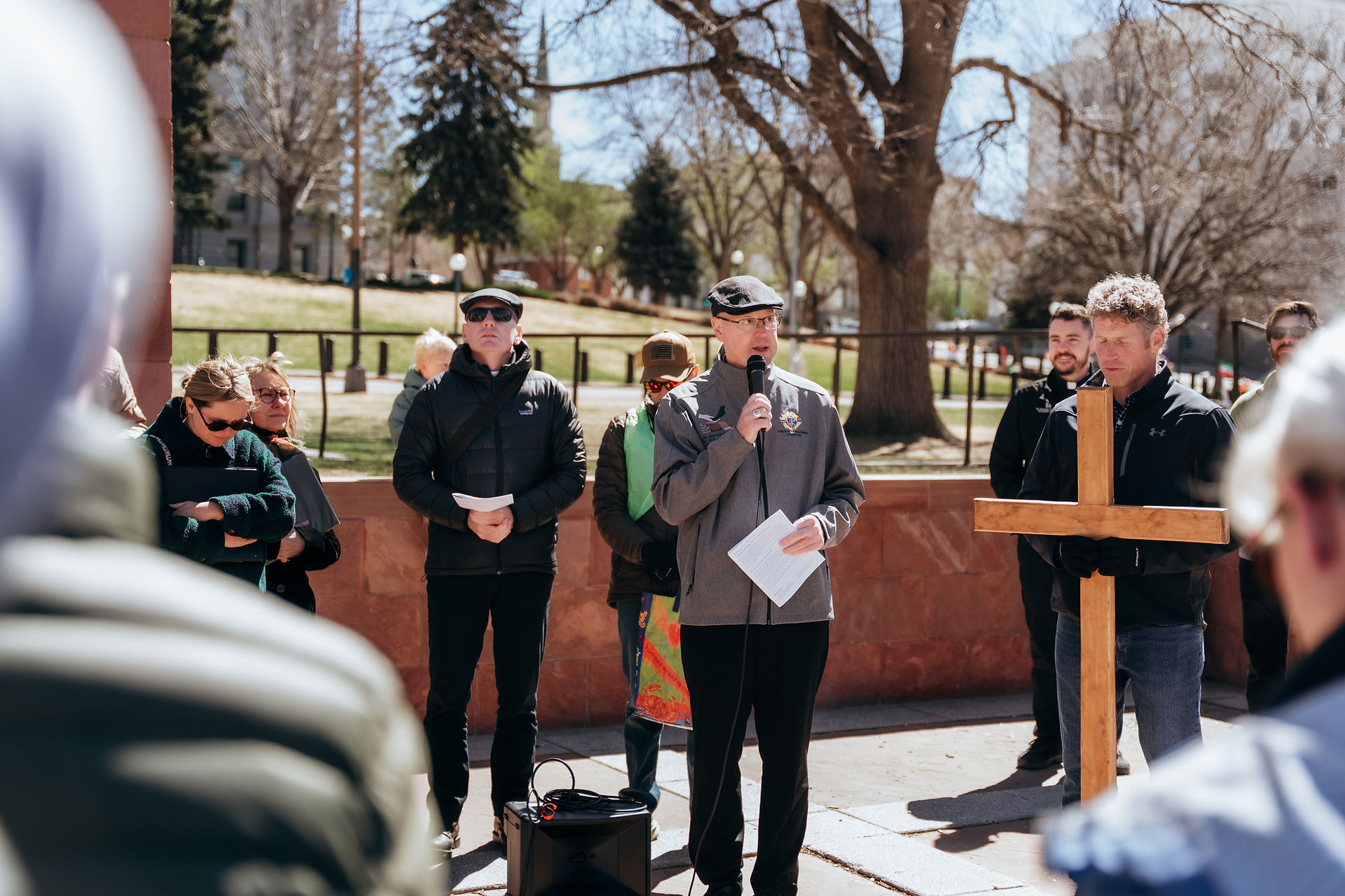 A man speaks into a microphone during an outdoor prayer gathering, standing near a wooden cross and a small crowd.