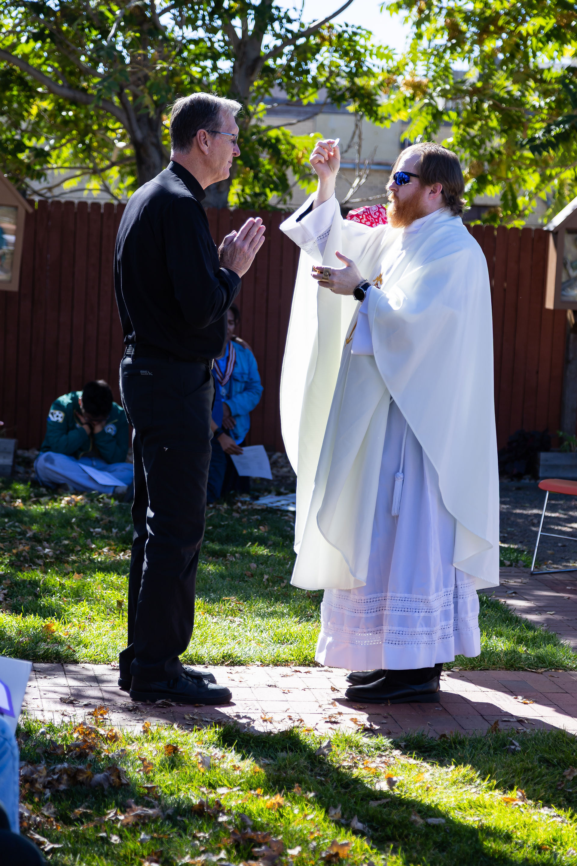 Priest in white vestment holds up Eucharist outdoors, facing man in black. Green grass, trees, and a wooden fence in the background.