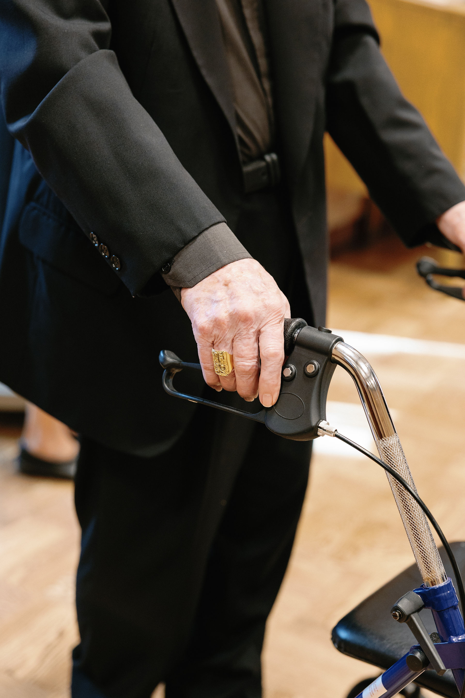 Elderly person in black suit holds a walker handle, wearing a gold ring. Background shows a wooden floor and blurred interior.