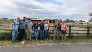 Smiling group of eight stands by a wooden fence at a farm, with barns, pens, and a cloudy sky behind them.