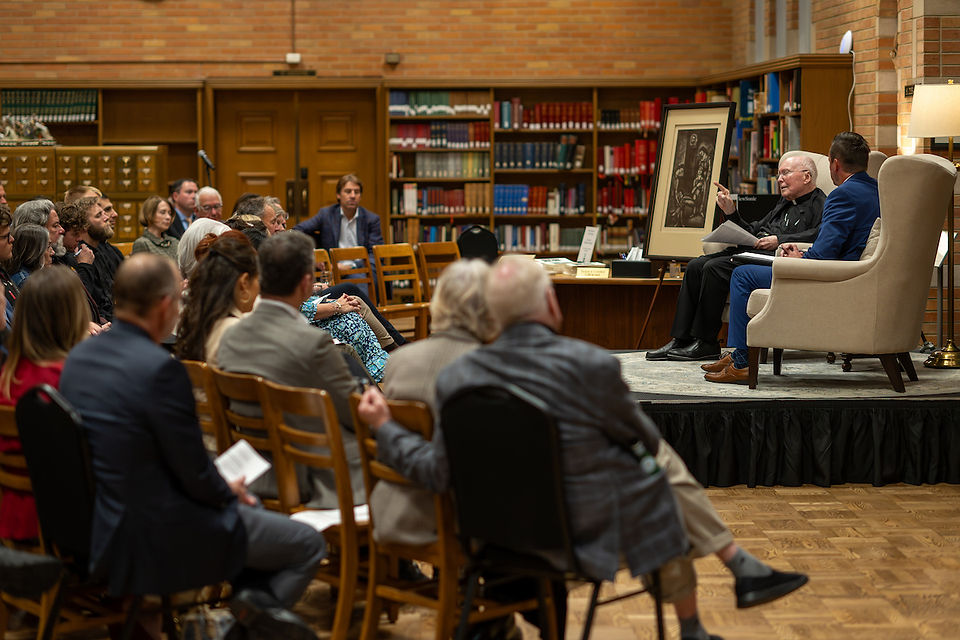 A speaker gestures at a library event with an audience seated on wooden chairs. Bookshelves and framed art are visible in the background.