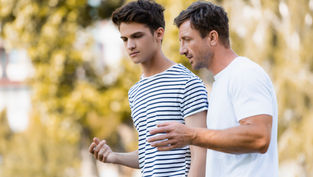 Two men walk outdoors, engaged in conversation. One wears a striped shirt, the other a white T-shirt. Trees are blurred in the background.