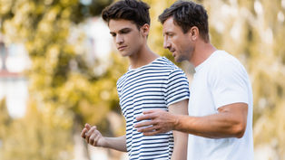 Two men walk outdoors, engaged in conversation. One wears a striped shirt, the other a white T-shirt. Trees are blurred in the background.