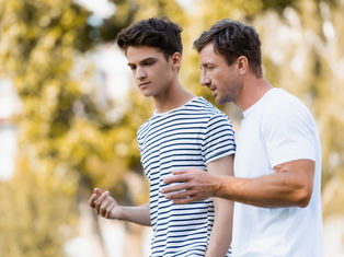 Two men walk outdoors, engaged in conversation. One wears a striped shirt, the other a white T-shirt. Trees are blurred in the background.