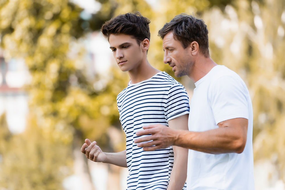 Two men walk outdoors, engaged in conversation. One wears a striped shirt, the other a white T-shirt. Trees are blurred in the background.