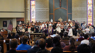 Church ceremony with clergy in purple robes and diverse group of people standing, stained glass windows behind, congregation seated.
