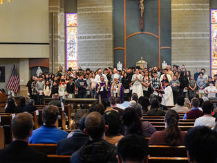 Church ceremony with clergy in purple robes and diverse group of people standing, stained glass windows behind, congregation seated.
