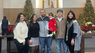 Family of six poses inside a decorated church with Christmas trees and wreaths. A child in red adds a festive touch. Smiling faces.