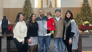 Family of six poses inside a decorated church with Christmas trees and wreaths. A child in red adds a festive touch. Smiling faces.