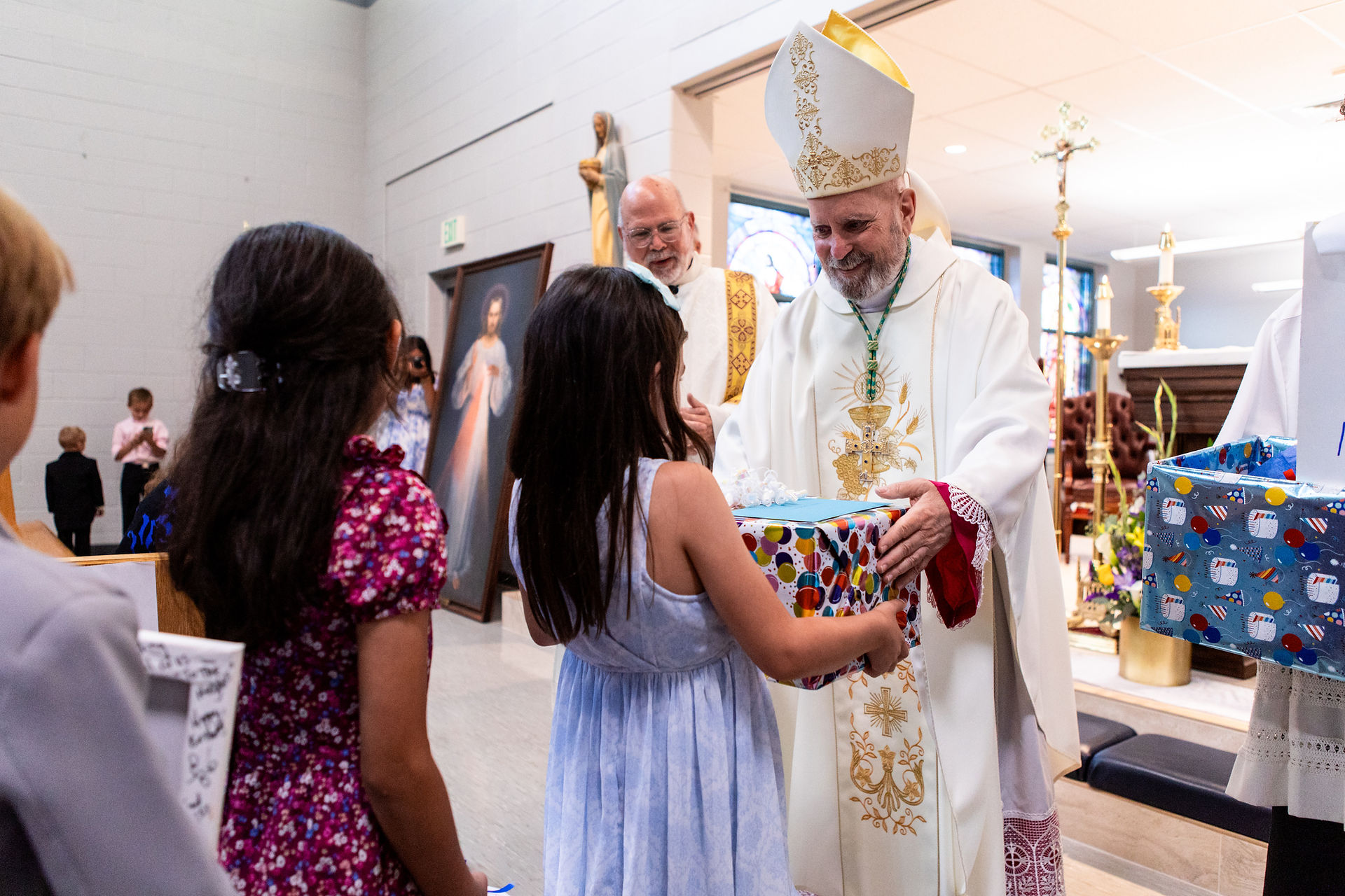 A bishop in white robes hands a gift to a girl in a church, with religious paintings and people observing in the background.
