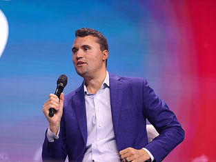 Man in a blue suit holding a microphone on stage, speaking. Background is a blurred gradient of blue and red, conveying a focused mood.
