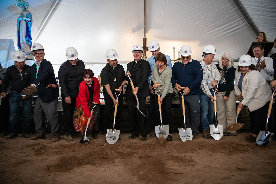 Clergy and community members wearing hard hats participate in a ceremonial groundbreaking with shovels.